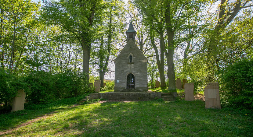 Josefskapelle - Eine kleine steinerne Kapelle steht inmitten einer grünen, von Bäumen umgebenen Lichtung, umgeben von einigen alten Grabsteinen.