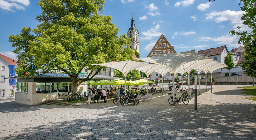 Ein Platz mit gepflastertem Boden und weißen Sonnenschirmen beherbergt ein Café mit Sitzbereichen unter einem großen Baum, umgeben von Fahrrädern, während im Hintergrund Gebäude und ein Kirchturm zu sehen sind.