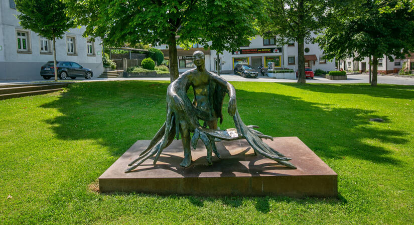 Schutzengel für Neuhausen auf dem Schlossplatz: Die Abbildung zeigt eine Skulptur auf einer Wiese, die eine menschliche Figur darstellt, deren Arme sich wie Äste ausbreiten.