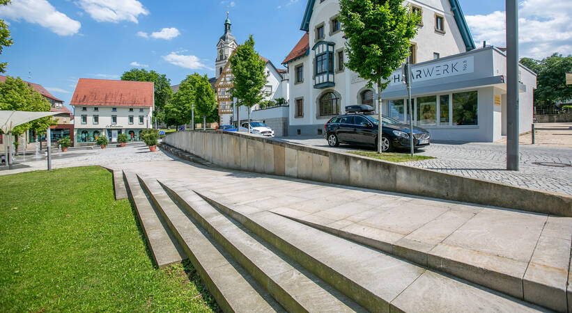 Schlossplatz Neuhausen: Eine Szene mit breiten Treppenstufen und einer Rampe, flankiert von Rasenflächen, führt zu einer gepflasterten Straße mit traditionellen Gebäuden und einem Kirchturm im Hintergrund.