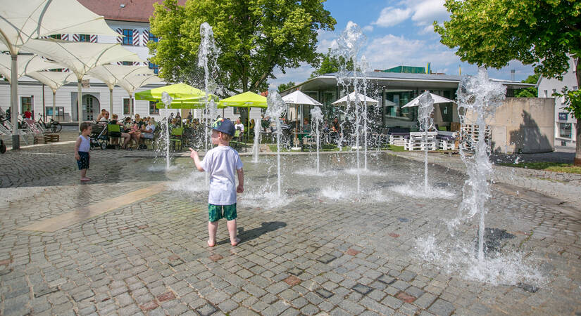 Ein kleiner Junge spielt bei sonnigem Wetter in einem Brunnen mit sprudelnden Wasserfontänen auf einem gepflasterten Platz, während Menschen in einem Café im Hintergrund sitzen.