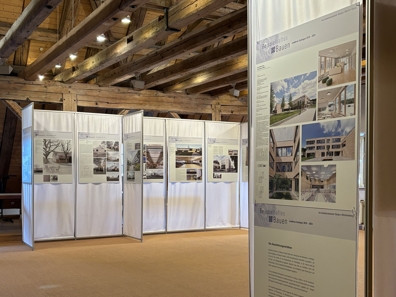 Eine Ausstellung in einem Holzgebäude zeigt mehrere Tafeln mit Architekturfotografien und Texten.