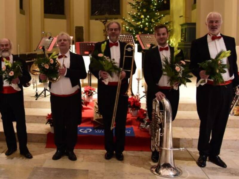 Fünf Männer in Anzügen stehen auf einer Bühne und halten Blumensträuße sowie Musikinstrumente, während im Hintergrund ein festlich beleuchteter Weihnachtsbaum zu sehen ist.